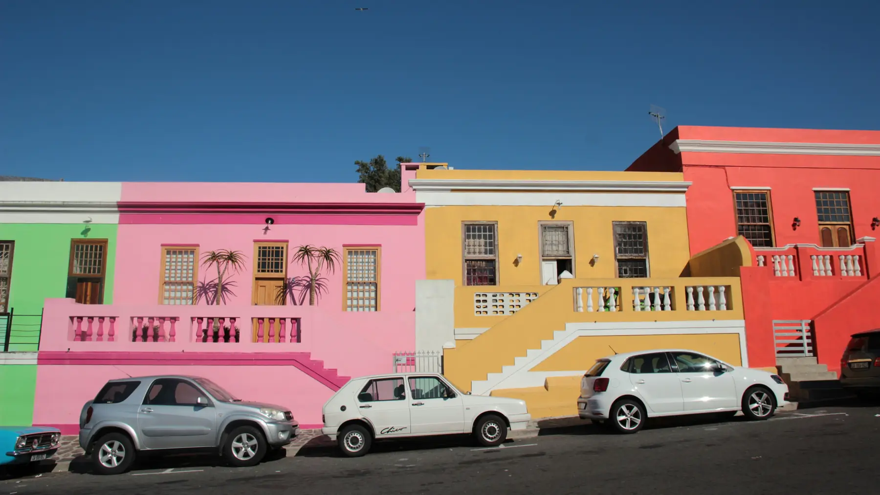 Brightly painted houses in green, pink, yellow and red along a street in the Bo-Kaap, Cape Town, South Africa, with cars parked in front.